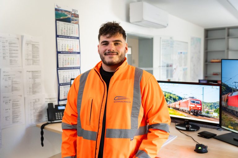 Ein Mann in orangefarbener Arbeitskleidung steht in einem Büro mit Monitoren im Hintergrund.