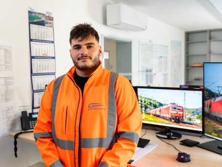 Ein junger Mann in orangefarbener Arbeitsjacke steht vor einem Schreibtisch mit Computern.