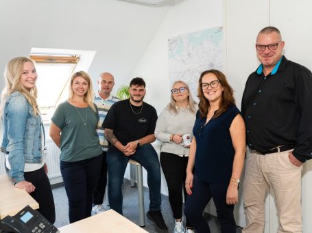 Sieben Personen in einem modernen Büro, lächelnd, stehen in einer gruppenfotoähnlichen Pose.